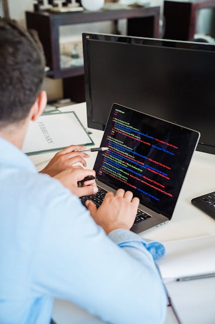 The image depicts a person seated at a desk in an indoor office environment, working on a laptop with a dark screen displaying colorful lines of code. The individual is using a mouse, with their back 